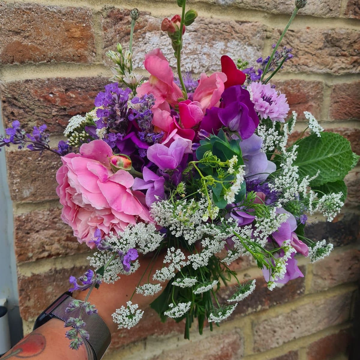 A hand-tied bouquet of sweet peas, roses and lavender against a brick wall